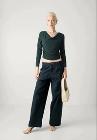 Top lungo a maniche lunghe verde con scollo a collo di cigno, jeans scuri a vita alta e gamba larga, e borsa beige. La modella indossa sandali con tacco trasparente.