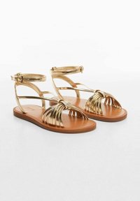 Zlaté metalické sandály s několika detaily pásků, s uzlovým designem na přední straně a hnědou plochou podrážkou. Nastavitelné spony na kotníku.