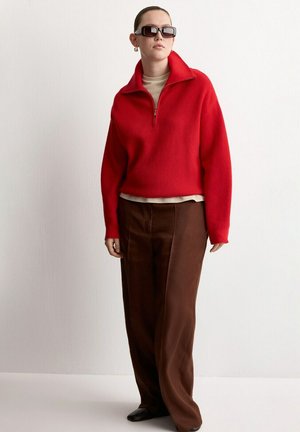 Mujer con gafas de sol oscuras, suéter rojo con cremallera, camisa beige, pantalones marrones holgados y zapatos negros, de pie contra una pared blanca lisa.