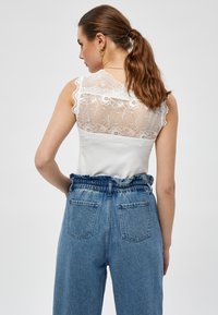 Top bianco con dettaglio in pizzo sulla schiena, bordi scallop, abbinato a jeans in denim elasticizzati a vita alta con una finitura blu sbiadita e tasche.