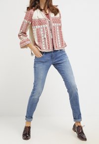 Chaqueta de patrón rojo y crema con mangas largas, combinada con jeans de mezclilla azules y zapatos de cordones marrones. Texturas y detalles sutiles evidentes.