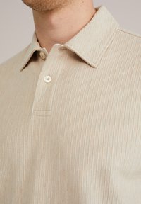 Bež majica s kratkimi rokavi polo sloga z teksturiranim, rebrastim vzorcem, ki ima koničast ovratnik in en ivorijn gumb.