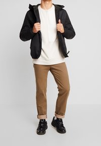 Black hooded jacket, white sweatshirt, brown pants, and black sneakers. Jacket has a zipper and elastic cuffs; pants are rolled at the hem.