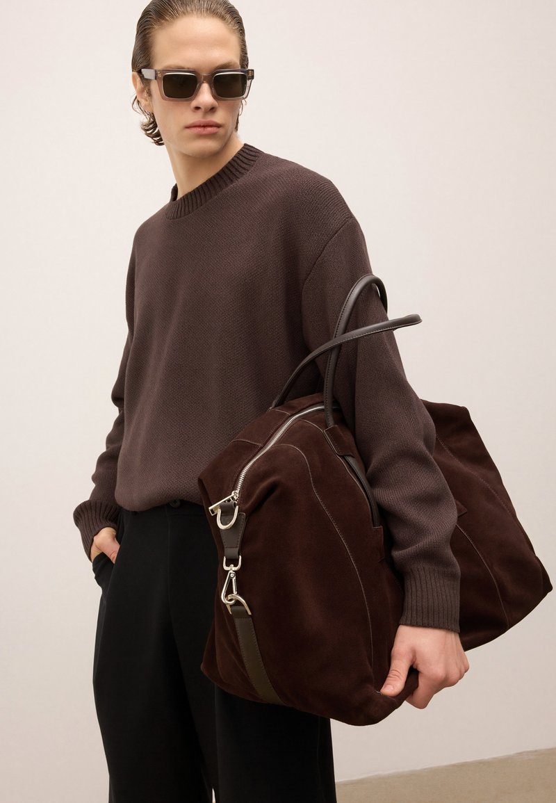 Ung person med slicket hår bærer mørke solbriller, en brun sweater, sorte bukser og en stor taske i mørkebrunt suede.