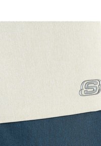 Bolso de lona con parte superior color crema y parte inferior azul. Presenta una tela texturizada y un acento de logo plateado en la parte frontal.