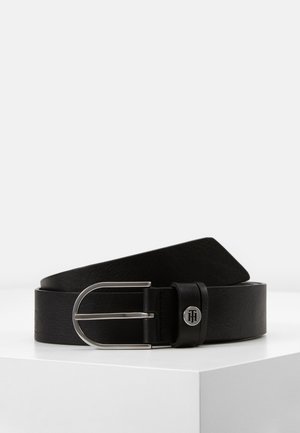 Zwarte leren riem met zilveren metalen gesp en klein rond logo-embleem, opgerold weergegeven op een witte ondergrond.