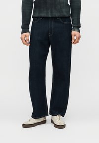 Άτομο που φοράει σκούρο μπλε τζιν με ίσια γραμμή, εκρού αθλητικά παπούτσια και σκούρο πράσινο πλεκτό πουλόβερ, στέκεται μπροστά σε λευκό φόντο.