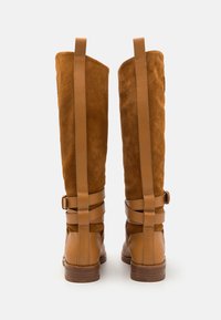 Bottes montantes en daim marron avec des sangles en cuir et des boucles, dotées d'un bout lisse et d'une tige texturée. Semelle en caoutchouc plate, hauteur du talon non visible.