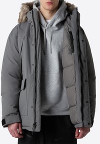 Grå isolerad jacka med furfoder på huvan, framsidan med dragkedja, tryckknappar och sidofickor, lagd över en grå hoodie.