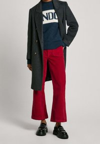 Donkergrijze dubbelgeknoopte jas over een marineblauwe trui met witte letters, gecombineerd met rode fluwelen uitlopende broek en zwarte loafers met studs.