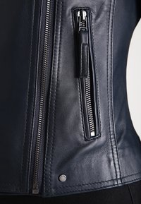 Námořnická kožená bunda s postranní kapsou na zip, štíhlým střihem a stříbrnými kovovými akcenty. Hladká textura a čisté švy.