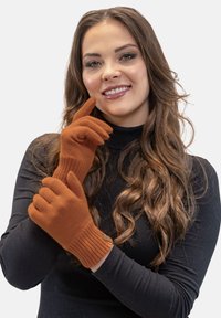 Brune strikkede handsker med ribbede manchetter, tekstureret overflade og en tætsiddende design. Modellen viser handskerne, mens hun har en sort bluse på.