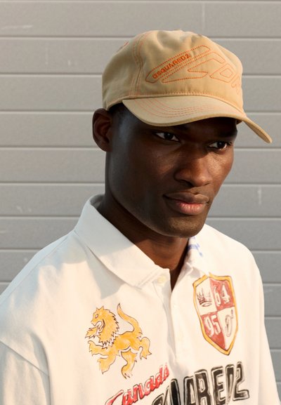 Casquette de baseball beige avec coutures et logo orange. Le mannequin porte un polo blanc avec un motif de lion jaune. En arrière-plan, un mur gris.