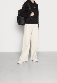 Person som bär en svart hoodie, vida vita byxor, vita sneakers och bär en svart ryggsäck på ena axeln mot en enkel bakgrund.