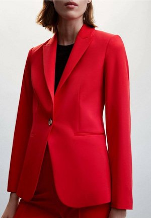 Femme portant un blazer rouge vif sur mesure à un seul bouton et un pantalon rouge assorti, avec un haut noir, sur un fond clair uni.