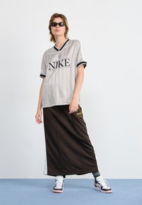 Maglia sportiva Nike grigia chiara con strisce nere e logo, abbinata a una lunga gonna marrone con tasche laterali e dettagli. Sneakers bianche ai piedi.