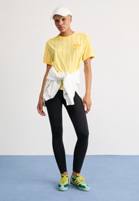 Person som bär en gul Adidas-tröja, svarta leggings, flerfärgade sneakers, en vit keps och en vit sweatshirt knuten runt midjan, stående på blått golv.