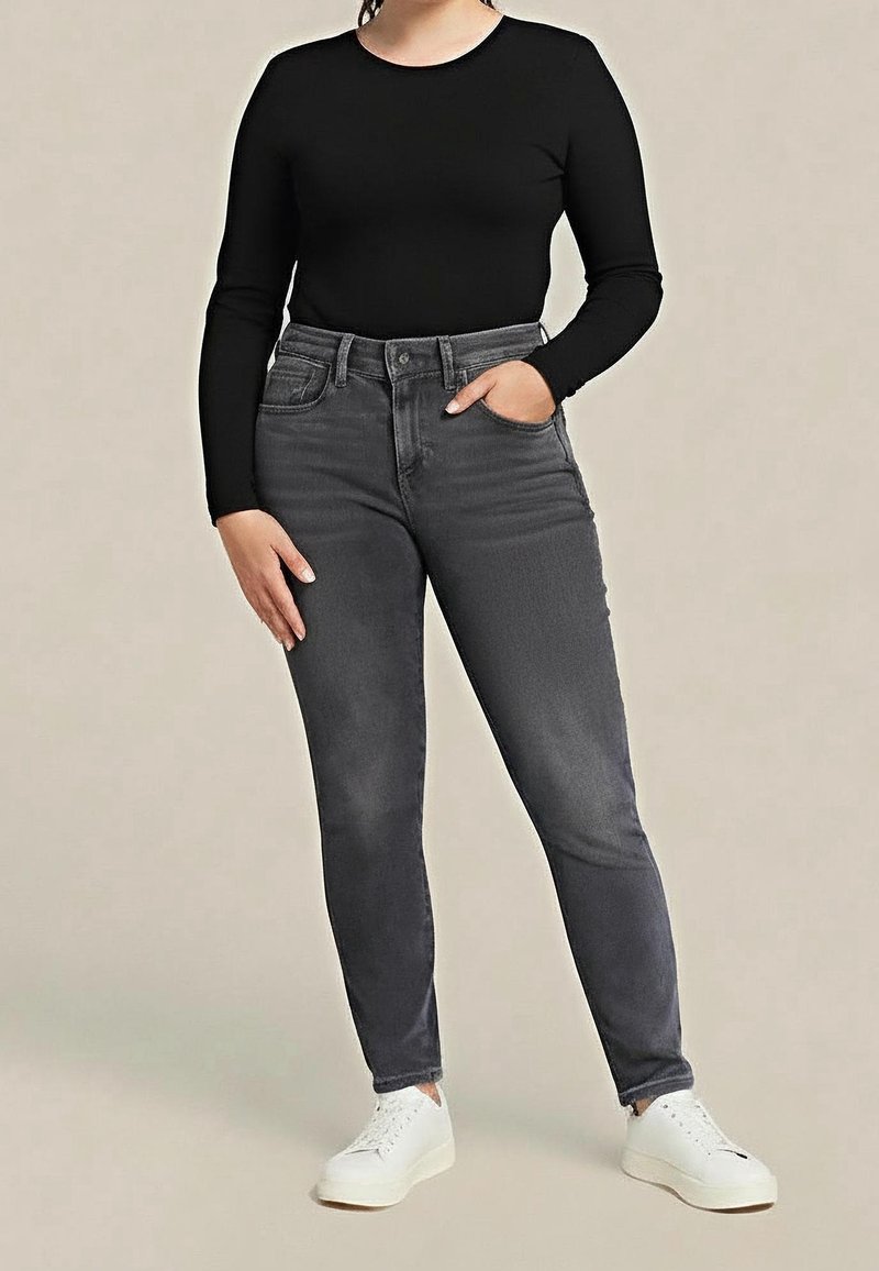 Personne portant un haut noir à manches longues, jean gris taille haute, baskets blanches, debout avec une main dans la poche devant un fond uni.