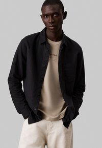 Camisa negra de botones sobre una camiseta beige, combinada con pantalones de color claro. Diseño simple con un bolsillo en el lado izquierdo de la camisa.