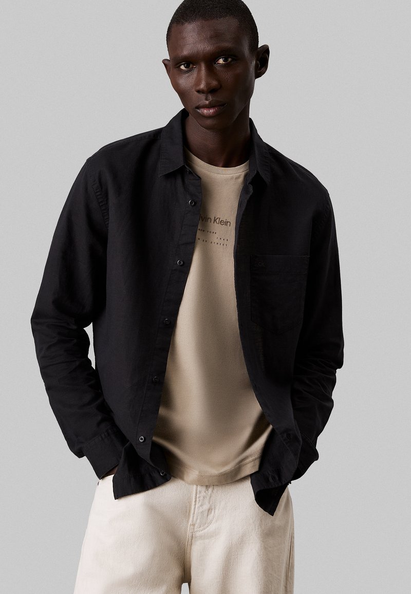 Camisa negra de botones sobre una camiseta beige, combinada con pantalones de color claro. Diseño simple con un bolsillo en el lado izquierdo de la camisa.