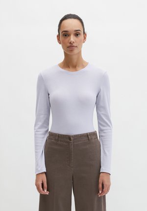 Woman with dark hair in a bun wearing a light purple long-sleeve top and high-waisted loose brown pants standing against white background.