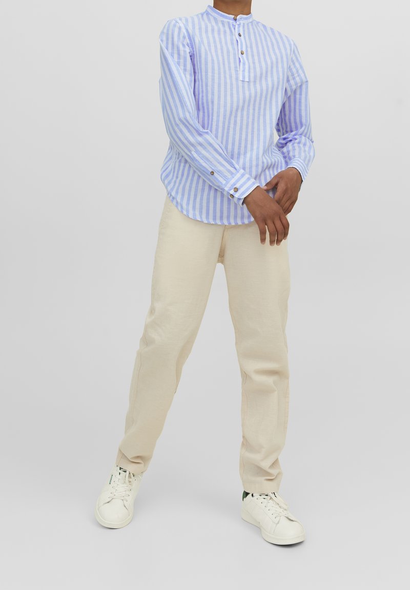Chemise à rayures bleu clair et blanche à manches longues avec un col haut et des détails de boutons, associée à un pantalon beige et des baskets blanches.