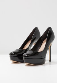 Chaussures à plateforme à talons hauts noires brillantes avec bouts arrondis, placées côte à côte sur une surface blanche devant un fond clair uni.