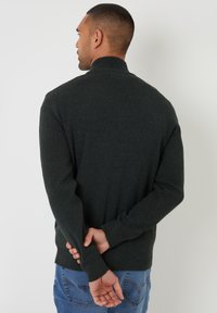 Donkergroen gebreid sweater met een hoge kraag en lange mouwen, met een gestructureerde afwerking. Gedragen met blauwe denim jeans, gezien vanuit de achterkant.
