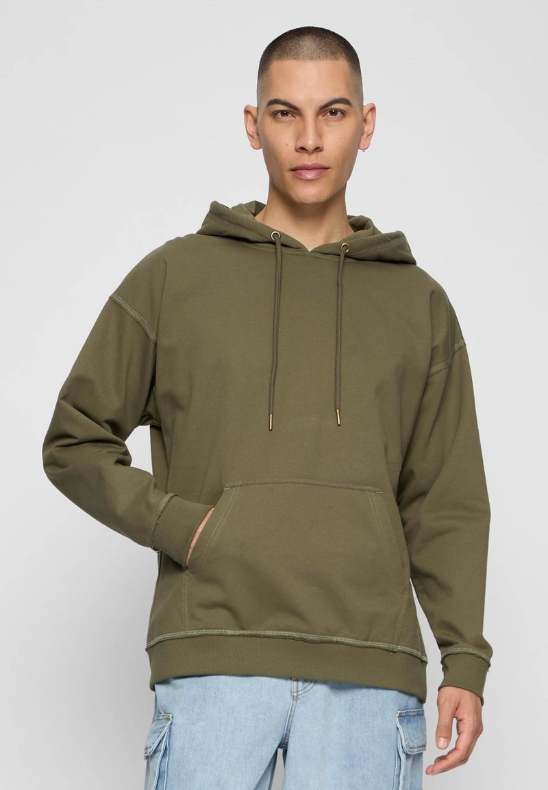 Olívazöld kapucnis pulóver puha, texturált anyagból, elülső zsebbel, húzózsinórokkal és kontrasztos varrási részletekkel. A modell világoskék cargo nadrágot visel.