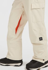 Beige skidbyxor med dragkedjefickor som har orange nätfoder, cargofickor, logotyget och en lös passform, matchade med svarta stövlar.