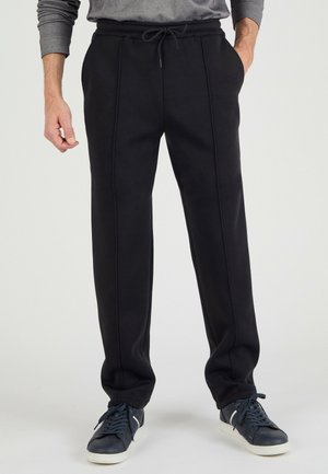 Man draagt zwarte broek met trekkoord en donkerblauwe sneakers, staat met één hand in de zak.