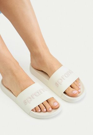 Stopy noszące białe klapki wsuwane z szerokimi paskami i wytłaczanym napisem, pokazujące starannie wykonany pedicure z jasnoróżowym lakierem do paznokci.