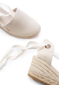 Espadrillas in tela beige con suola in corda di juta e lacci alla caviglia. Presentano un tacco a motivo a scacchi e un dettaglio del logo dorato.