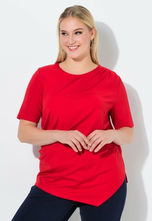 Smilende kvinde med langt blondt hår iført øreringe, rød kortærmet skjorte med asymmetrisk kant og navy bukser, stående mod en hvid væg.