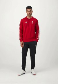 Sarkans sporta krekls ar baltām svītrām un logo, apvienots ar melniem sporta biksēm. Baltas sporta kurpes ar sarkaniem un zilajiem akcentiem.