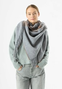Grauer Schal mit Fransen und subtilen Farbverläufen, über einem hellblauen Pullover getragen. Besticktes Blumendetail in der Ecke.