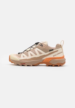 Chaussure de randonnée marron clair et crème avec semelle orange, système de laçage rapide, étiquette Gore-Tex et logo Salomon sur le côté.