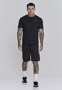 Musta puuvillapaita, jossa on pieni logo, yhdistetty mustiin shortseihin, joissa on sivutaskut. Asusteena valkoiset Nike-sukat ja mustat lenkkarit.