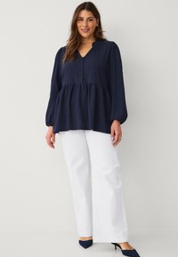 Top peplum azul marino con escote en V, detalles de botones y mangas largas abullonadas. Combinado con pantalones anchos blancos y tacones azul marino.