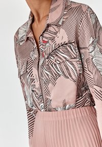 Blusa rosa con stampa botanica in bianco e nero, dotata di colletto, due tasche anteriori e gonna plissè rosa con texture.