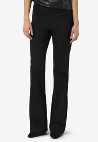 Osoba u crnim širokim hlačama i crnim cipelama s oštrim vrhom, stoji nasuprot bijeloj pozadini.