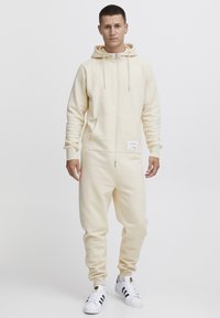 Salopette color crema con zip frontale, tasche grandi, cappuccio e polsini a coste. Presenta un'etichetta del marchio vicino alla vita. Indossata con sneakers bianche.