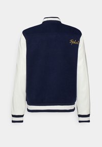 Marineblauwe en witte varsityjack met geribbelde kraag en zoom, voorzien van geborduurd "Rebel" in goud op de achterkant. Gladde textuur, contrasterende mouwen.