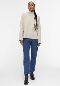 Beige høyhalset genser i et mykt, strukturert strikket materiale, parret med rette blå jeans og svarte ankelstøvler. Enkel, moderne design.
