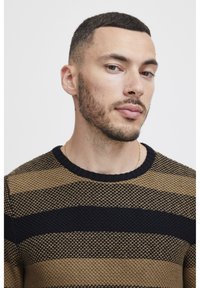 Maglione a righe con tonalità nere e marroni, caratterizzato da un motivo a maglia testurizzata e un collo a costine. È visibile un accento in catena dorata.