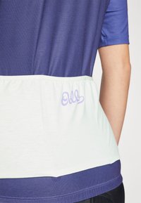 Kortärmad topp med en mörkblå övre del och en ljusgrön nedre del, med rynkad tyg och lila "Odds" logotyp på baksidan.