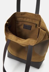 Braune Canvas-Tote-Tasche mit dunklen Griffen, die eine Reißverschlusstasche im Inneren und einen flachen Boden für Stabilität aufweist. Sichtbares Lederakzentetikett.
