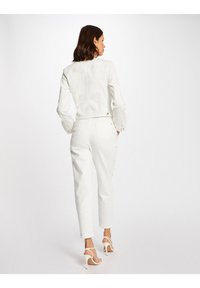 Giacca in denim bianco con design cropped, abbinata a pantaloni bianchi. Le caratteristiche includono maniche arrotolate e una silhouette elegante. Tacchi a spillo in primo piano.