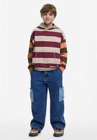 Stribet hættetrøje i beige, bordeaux, grøn og orange; kængurulomme; blå løse cargobukser med lyseblå patches; brune sneakers.