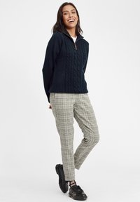 Pullover blu navy a maglia cable-knit con mezzo zip e cerniera beige, abbinato a pantaloni grigi a quadri affusolati e scarpe nere.